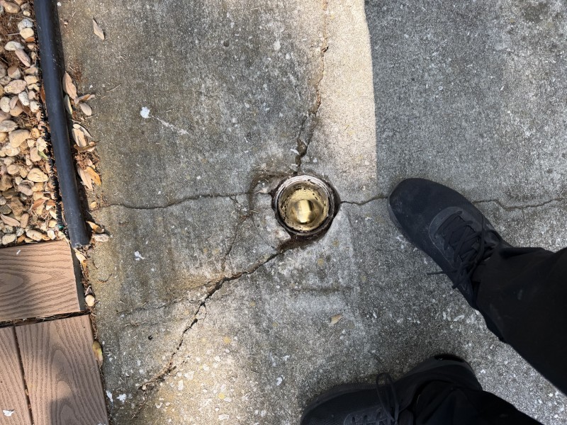 Ground-level view of a PVC sewer cleanout cap set flush in concrete pavement with a plumber boot visible beside it, South Tampa FL, April 2026