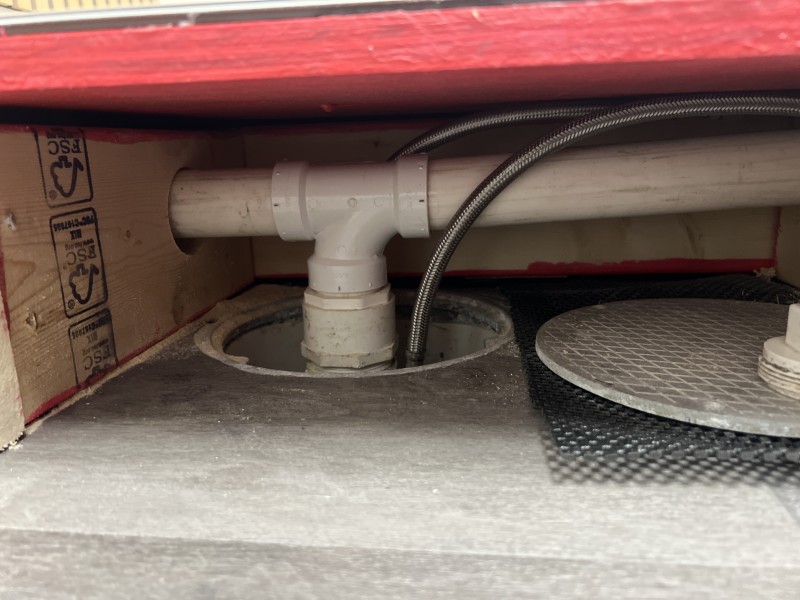 Side view under pedicure platform showing white PVC tee drain fitting connected through the platform floor with braided stainless steel supply lines running alongside, Tampa FL, May 2026