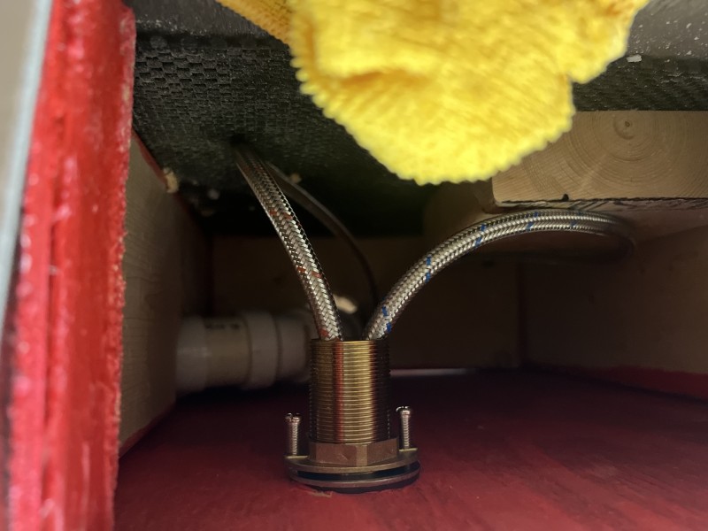 Under-platform close-up of brass faucet deck mount with two braided stainless steel hot and cold supply hoses connected through the platform surface, white PVC drain visible in background, Tampa FL, May 2026
